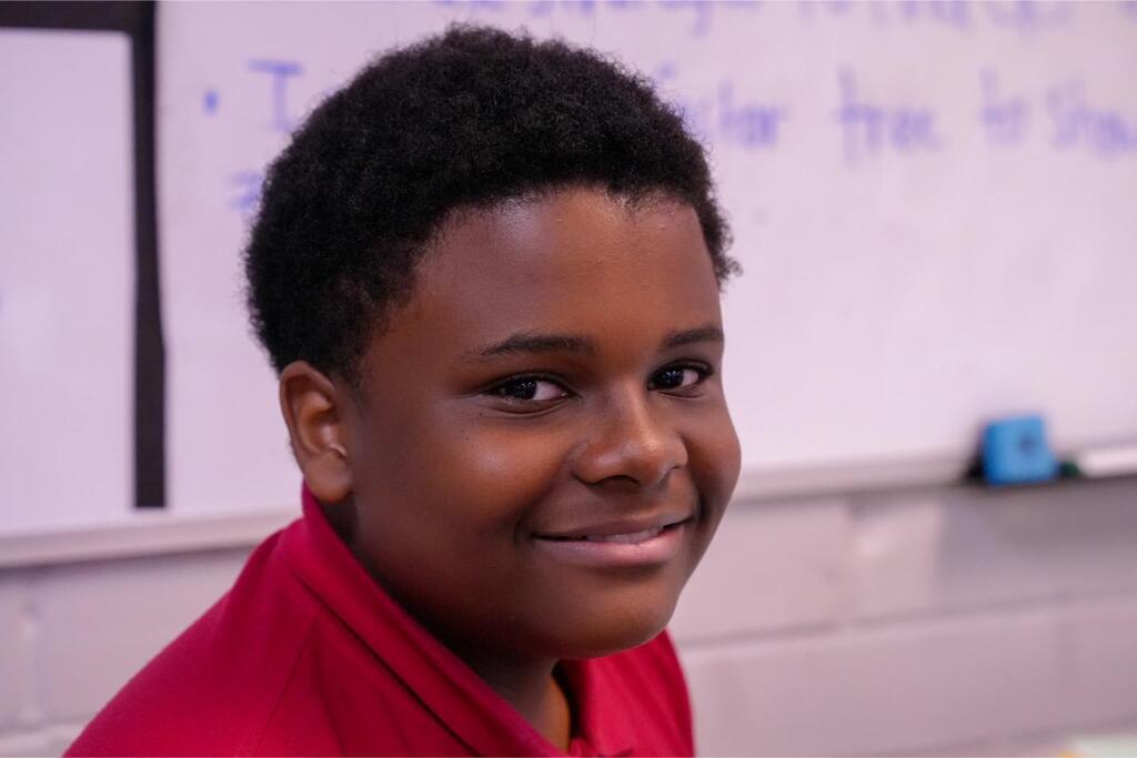 A smiling young person in a red shirt stands in front of a whiteboard in a classroom.