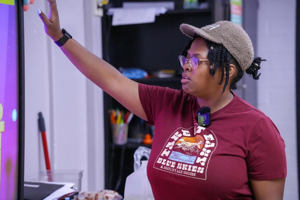 A woman in a maroon t-shirt and cap points at a screen. Behind her is a bookshelf with books and pens.