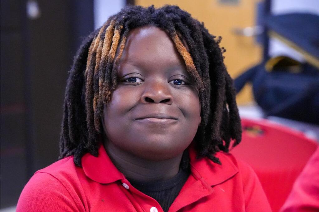 A person with braided hair wearing a red shirt, sitting against a blurred background.