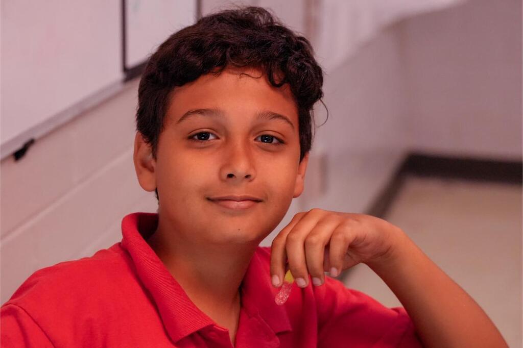 A young boy in a red shirt sits at a desk with a smile, one hand on his chin.