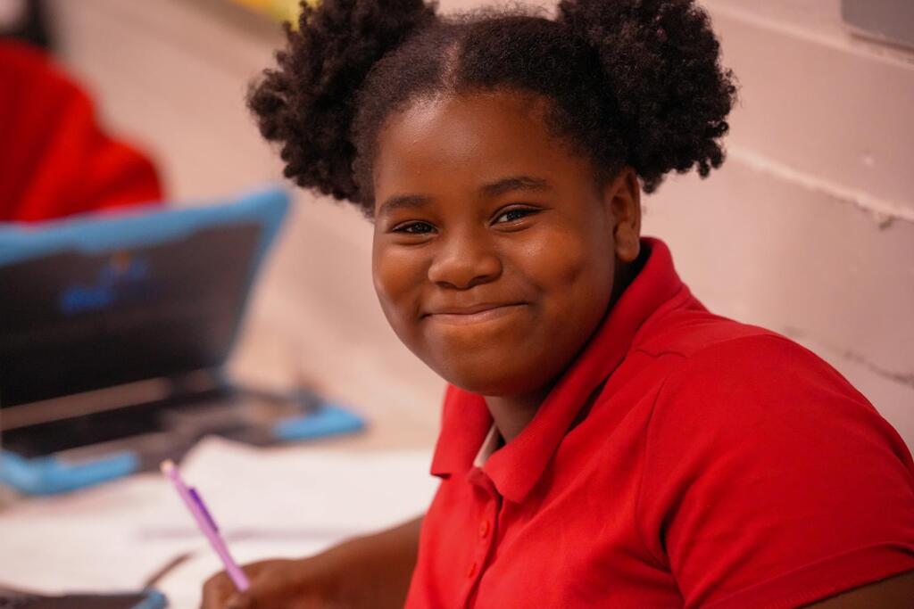 Girl in red shirt seated at desk with laptop. She holds a pen and smiles.