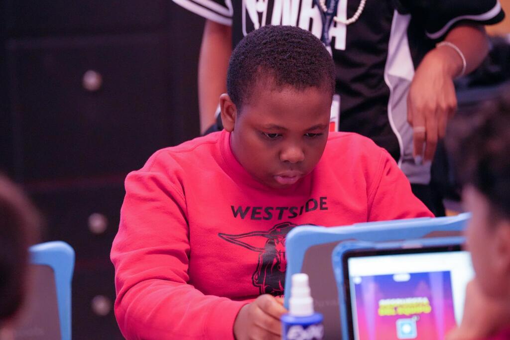 A child sits at a table with a laptop, focused on the screen. Behind them, a person in a black and white jersey stands.