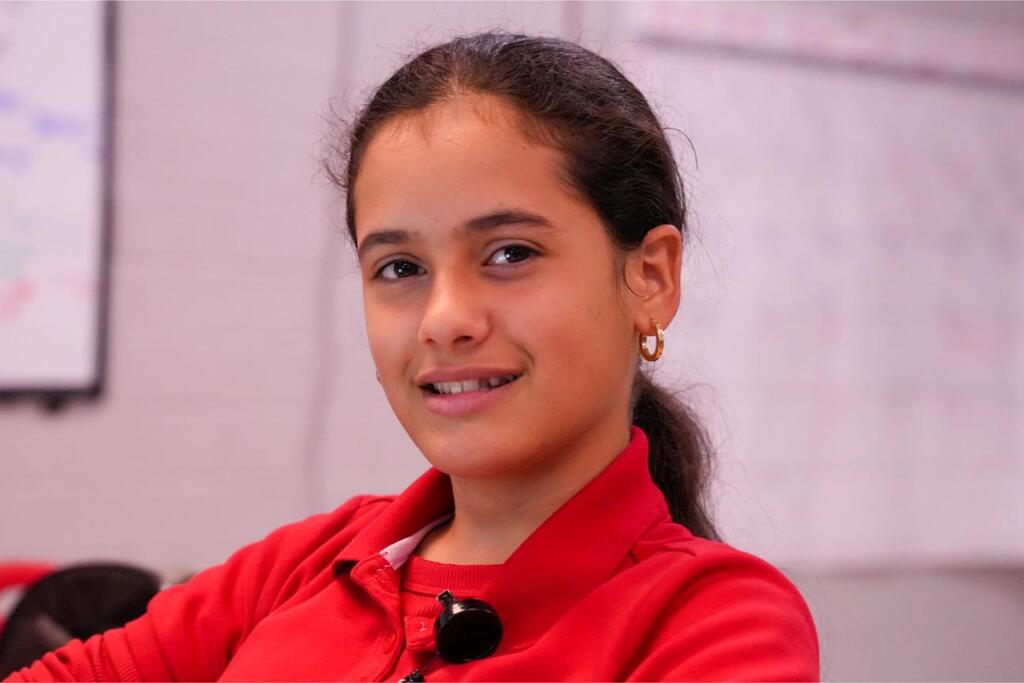 A young girl wearing a red shirt smiles at the camera. She has dark hair and is seated indoors.