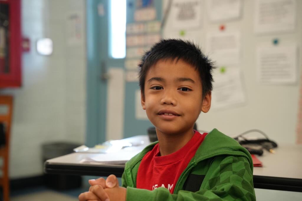 Child in green jacket smiles in classroom, hands clasped. Behind, a white wall with papers and a door.