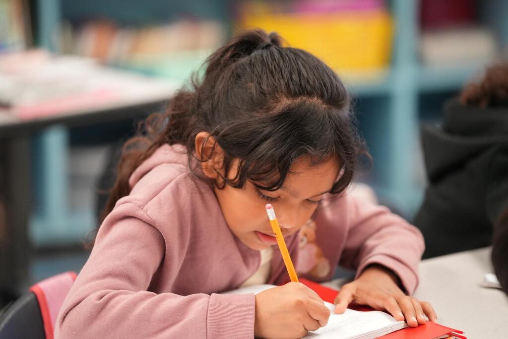 A girl in a pink shirt focuses on writing on paper with a pencil in a classroom.