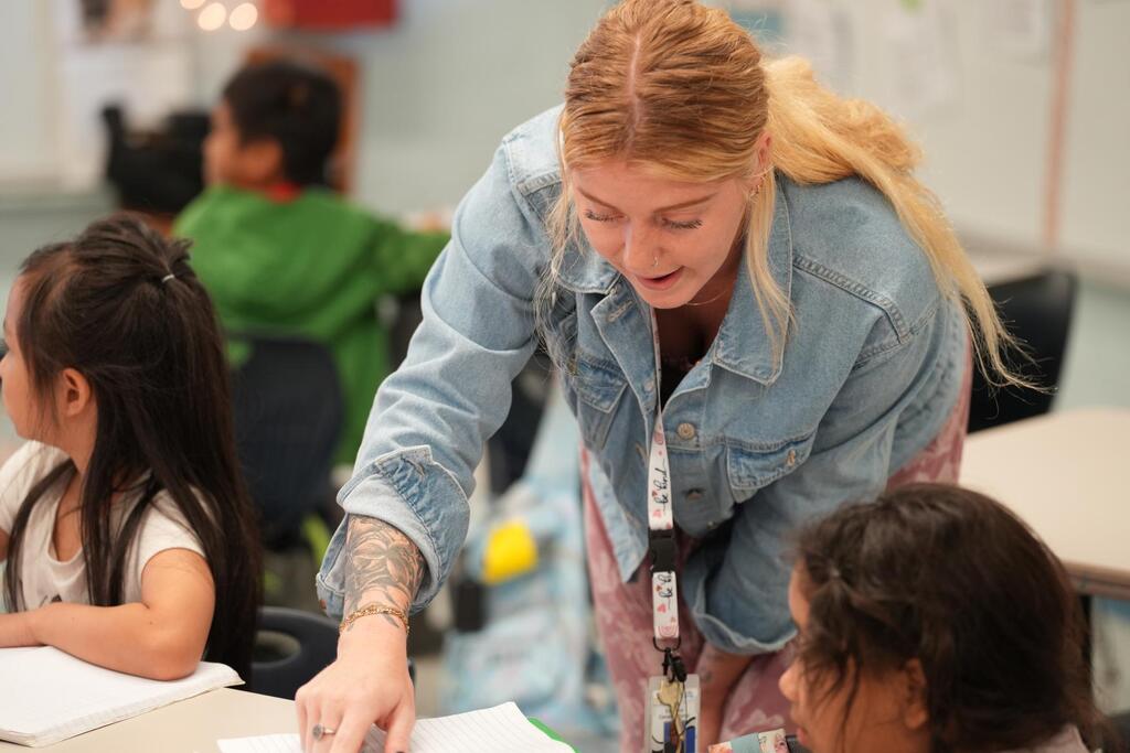 Teacher with a ponytail assists a student with a paper at a desk. Background shows a classroom.