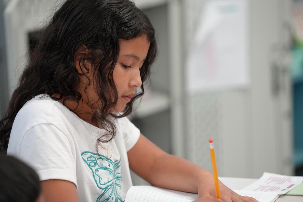 A young girl sits at a table, focused on writing in a book with a yellow pencil.