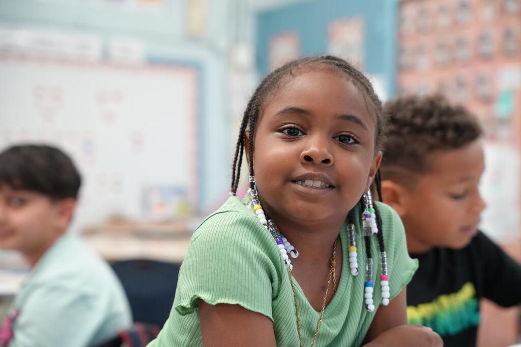 A girl with braids in a green shirt sits in a classroom, looking at the camera. Behind her, two children sit at desks.
