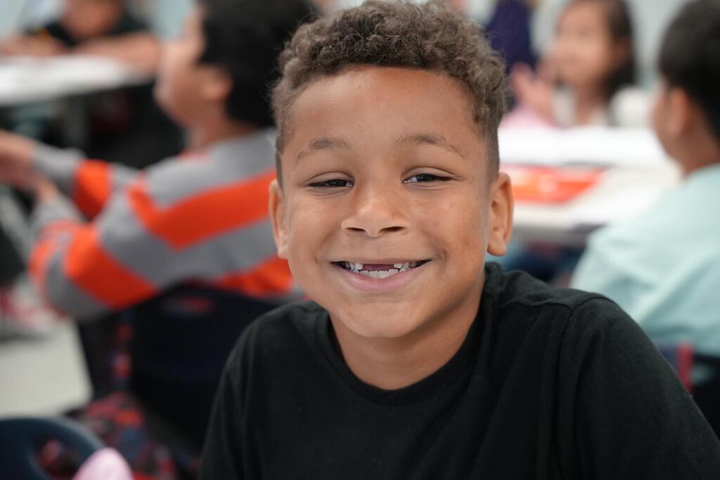 A boy with a big smile sits at a table among other students. The background shows other students seated at desks.