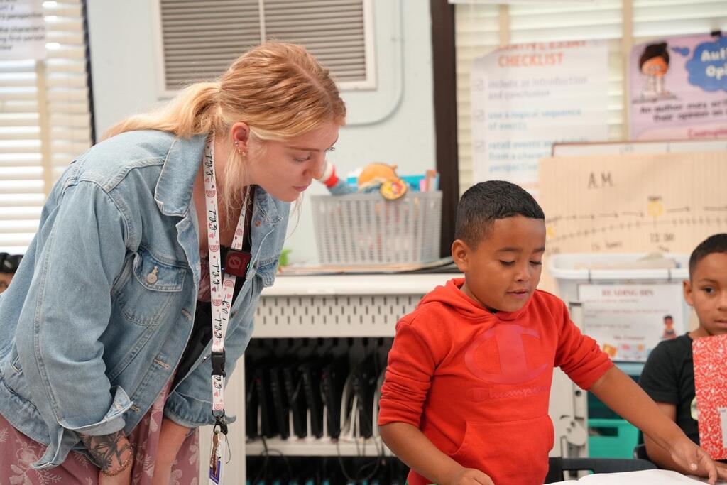 A woman helps a boy at a desk while another boy watches. A bulletin board and books are behind them.