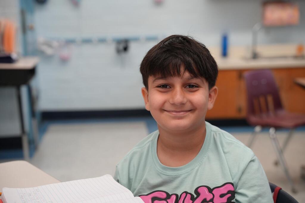 A boy in a gray shirt with pink letters sits at a table holding paper, smiling.