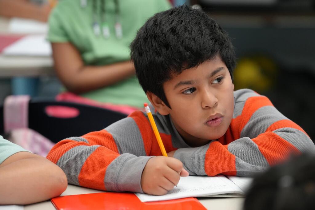 A young boy writes on a piece of paper in a classroom. He wears an orange and gray long-sleeve shirt.