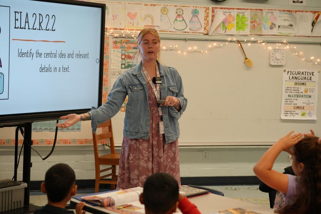 A teacher explains a lesson with a screen displaying "ELA2R22" behind her. Children sit attentively, with one raising a hand.
