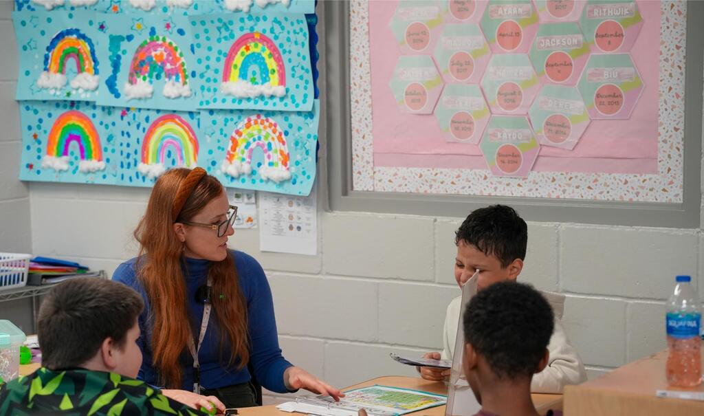 Woman in glasses speaks with four children at a desk. Rainbow posters on the wall behind them.
