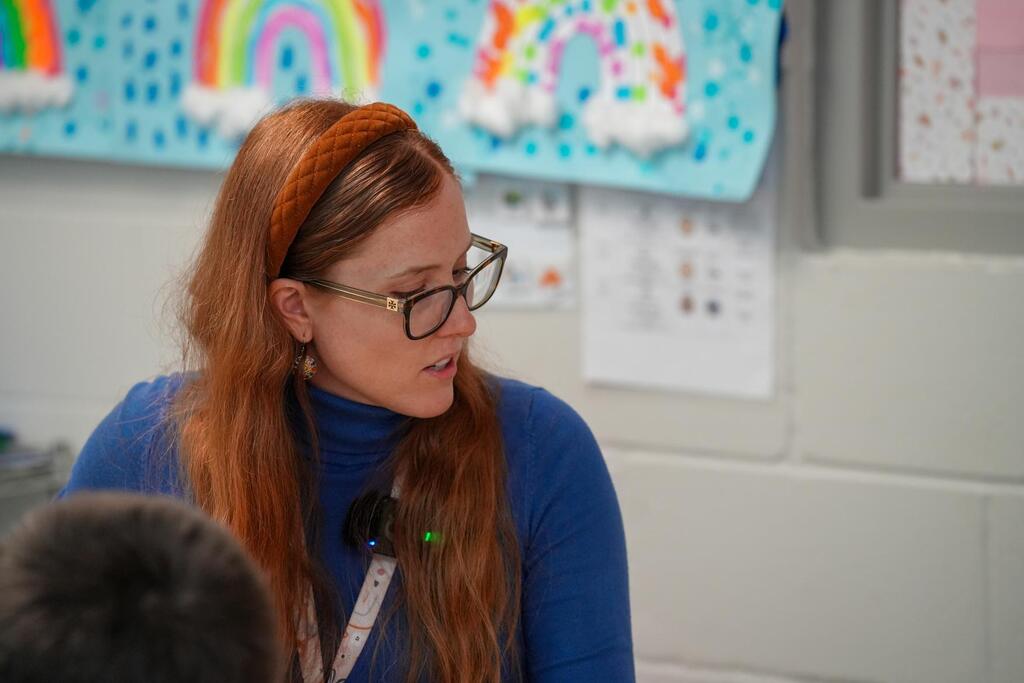 Woman with red hair and glasses speaks to student. Background includes bulletin board with rainbow and clouds.