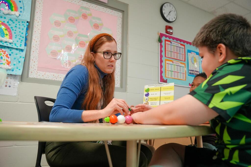 Woman and child at a table, woman holding multi-colored beads. Classroom background with bulletin board and clock.
