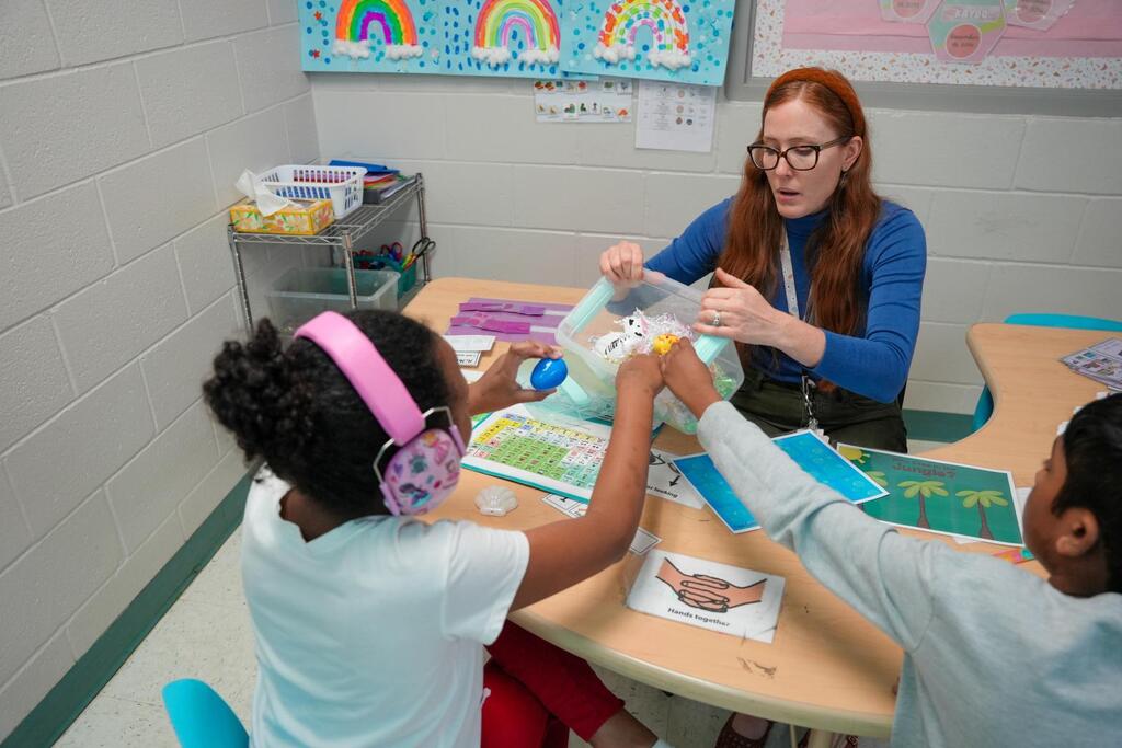A woman in glasses assists two children wearing headphones, one holding a stuffed animal, seated at a desk.