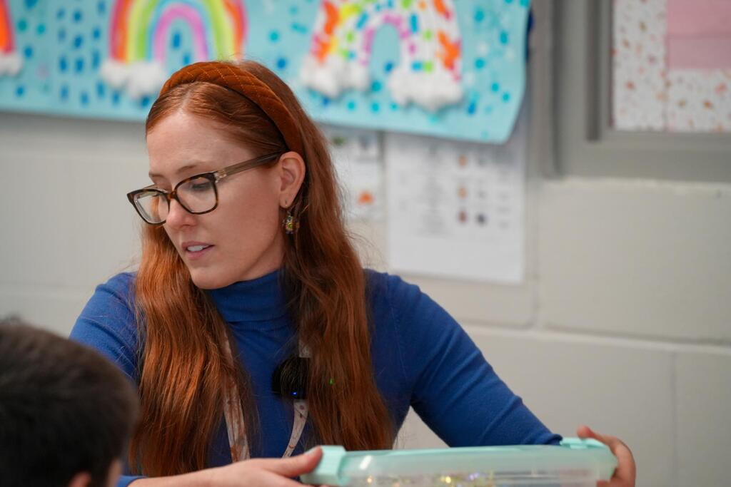 Woman with long red hair wearing glasses leans over and shows a clear container to a student.