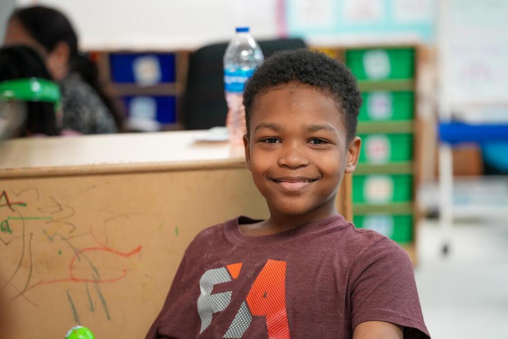 A young boy with a smile poses in a classroom. A counter with a water bottle and storage boxes behind him.