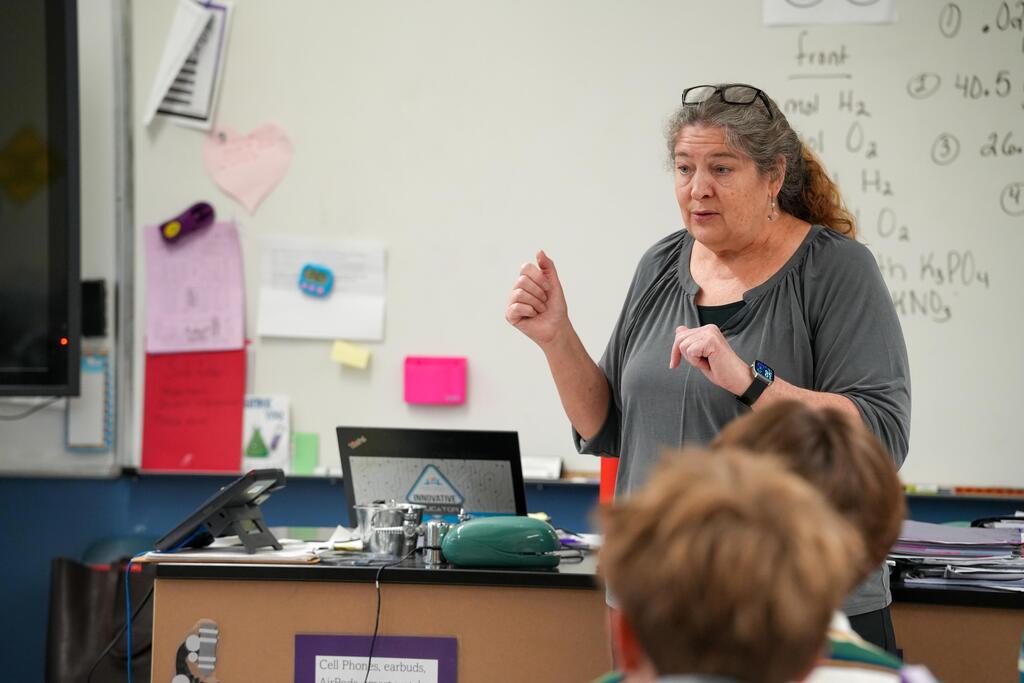 A woman speaks to students in a classroom. She stands at a desk with a laptop. Behind her, a whiteboard displays equations.