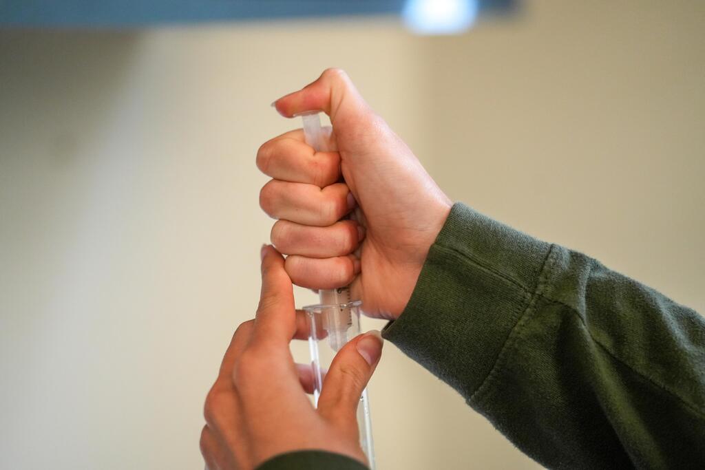 A person in a green shirt holds a transparent plastic vial with their thumb and index finger.