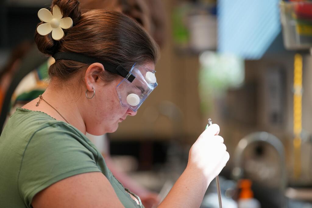 A woman in a green shirt and white goggles holds a tool. Behind her, a blurry background shows other objects.