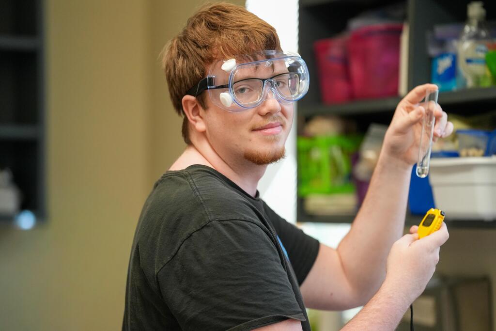 Person in a black shirt and safety glasses holding a pipette and test tube indoors.