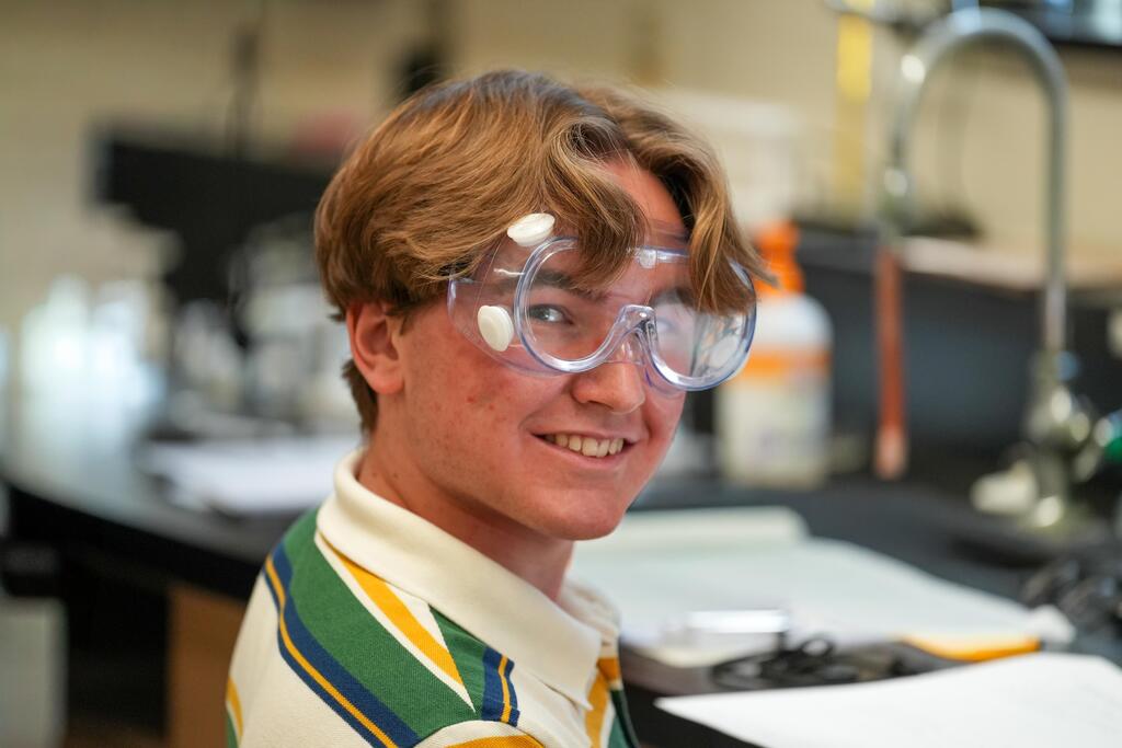 A person in goggles smiles at the camera in a lab. Behind them, lab equipment and supplies are on the counter.