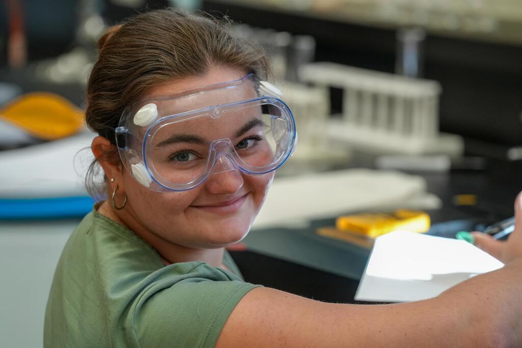 A woman in a green shirt and safety goggles, smiling while writing on a piece of paper.