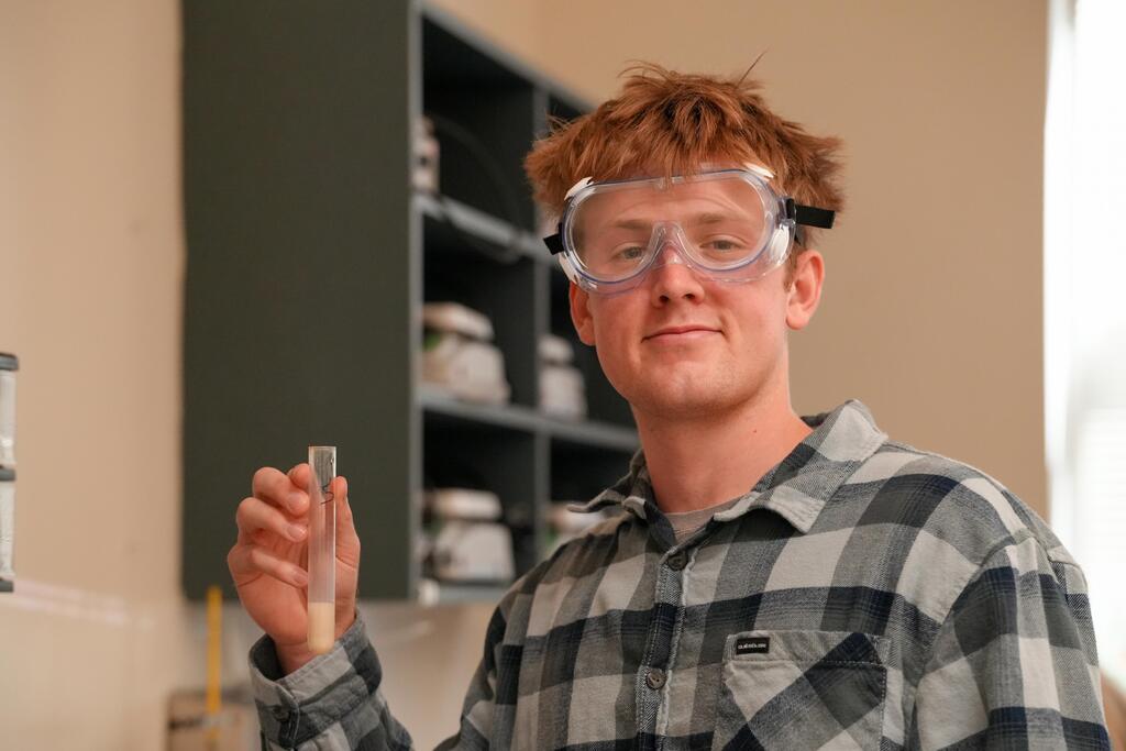 A person in a plaid shirt and goggles holds a test tube inside a room with shelves.