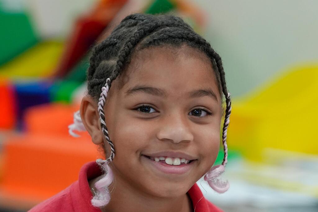 A young girl with braided hair and white hair ties smiles. She has a red top. Behind her, colorful shapes of toys are blurred.