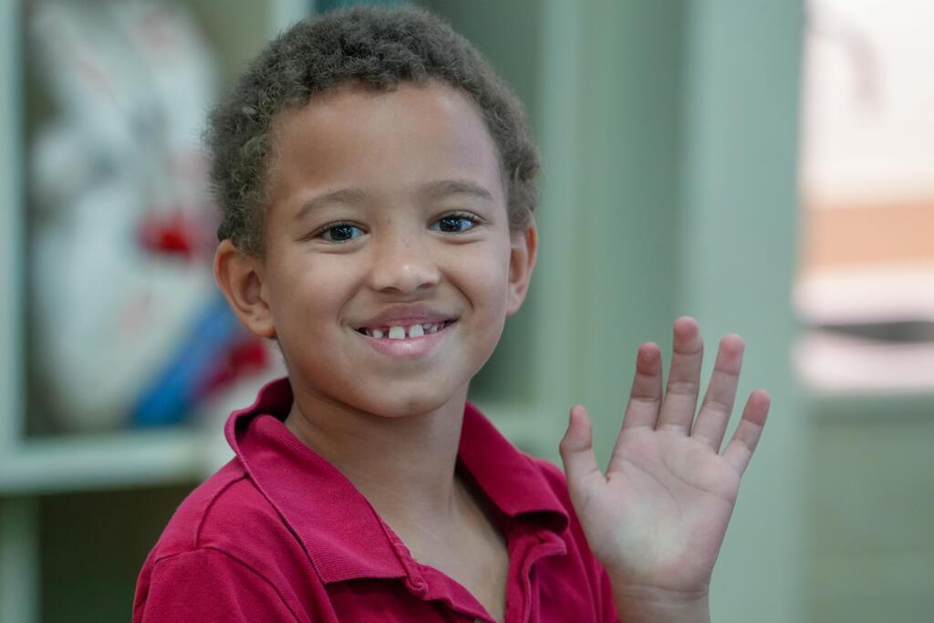 A child smiles and waves with a red shirt, standing near a green shelf with toys.