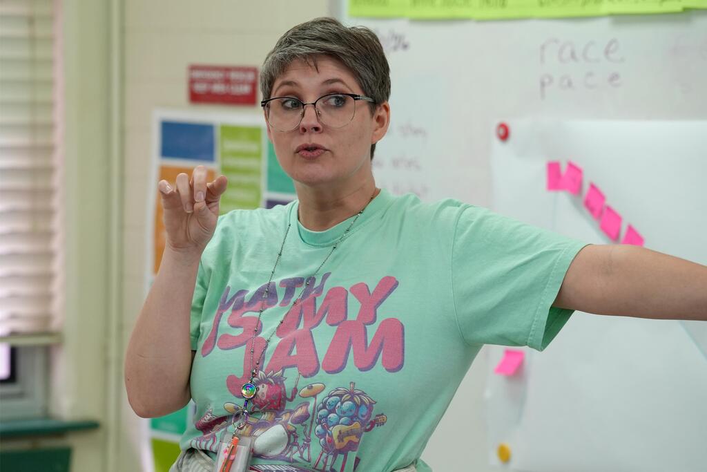 Woman wearing glasses and a green shirt with "Is My Jam" on it speaks with arms extended.