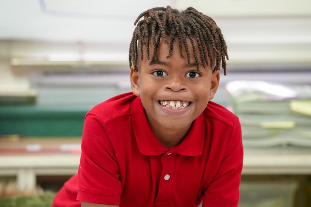 A young child with braided hair, wearing a red polo shirt, smiles and shows teeth. Background is blurred.