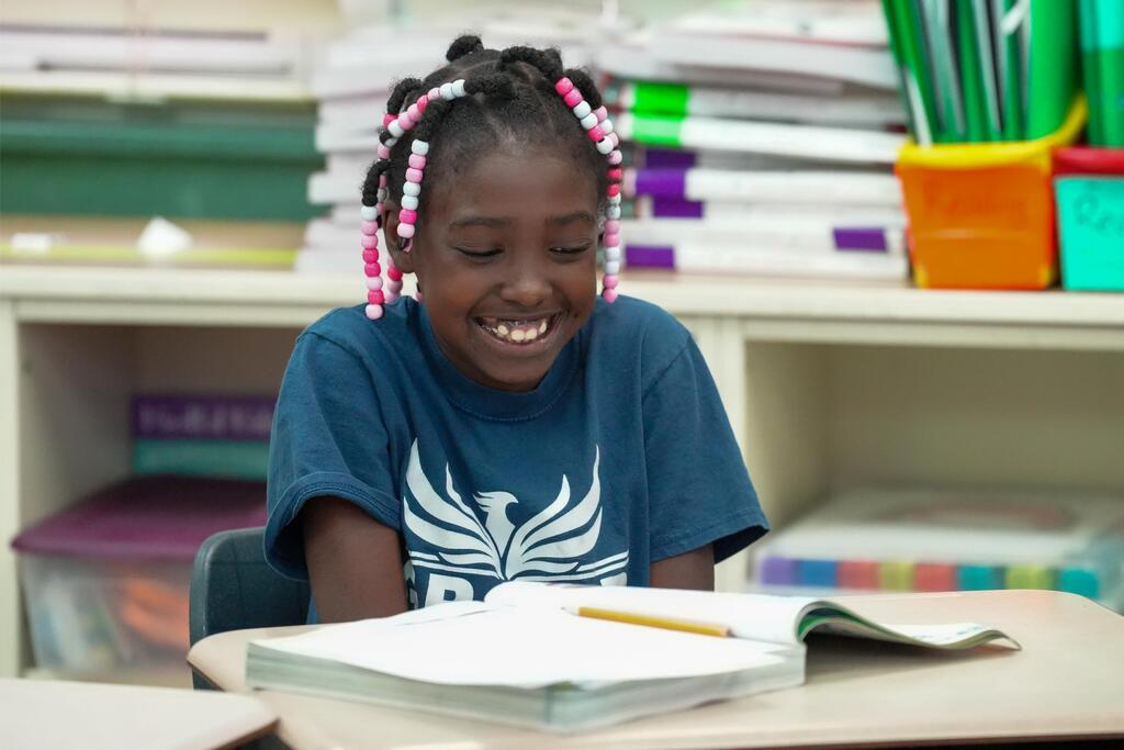 A young girl in a blue shirt sits at a desk, smiling and reading a book.
