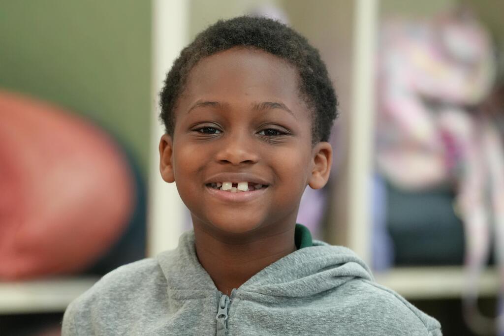 A young boy in a gray hoodie smiles, standing against a backdrop of a white shelving unit.