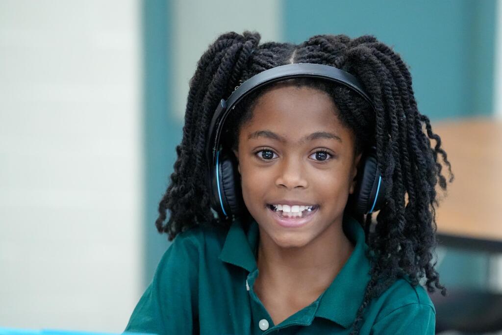 A young girl with braided hair, wearing headphones, smiles brightly. She sits in a room with blue walls and white trim.