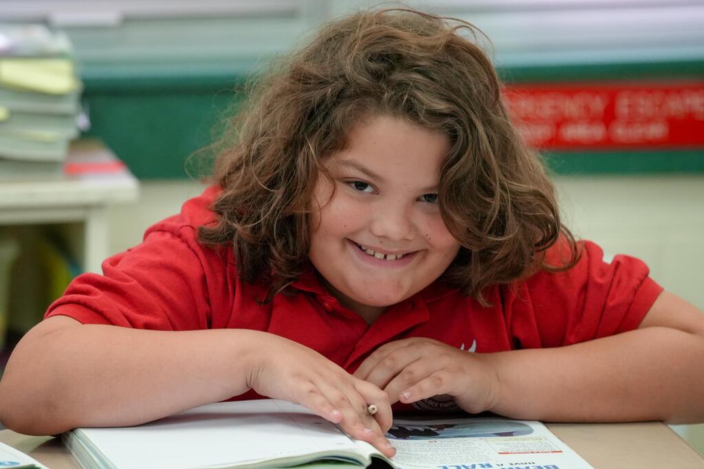 A young girl with curly hair smiles while seated at a desk, holding an open book.