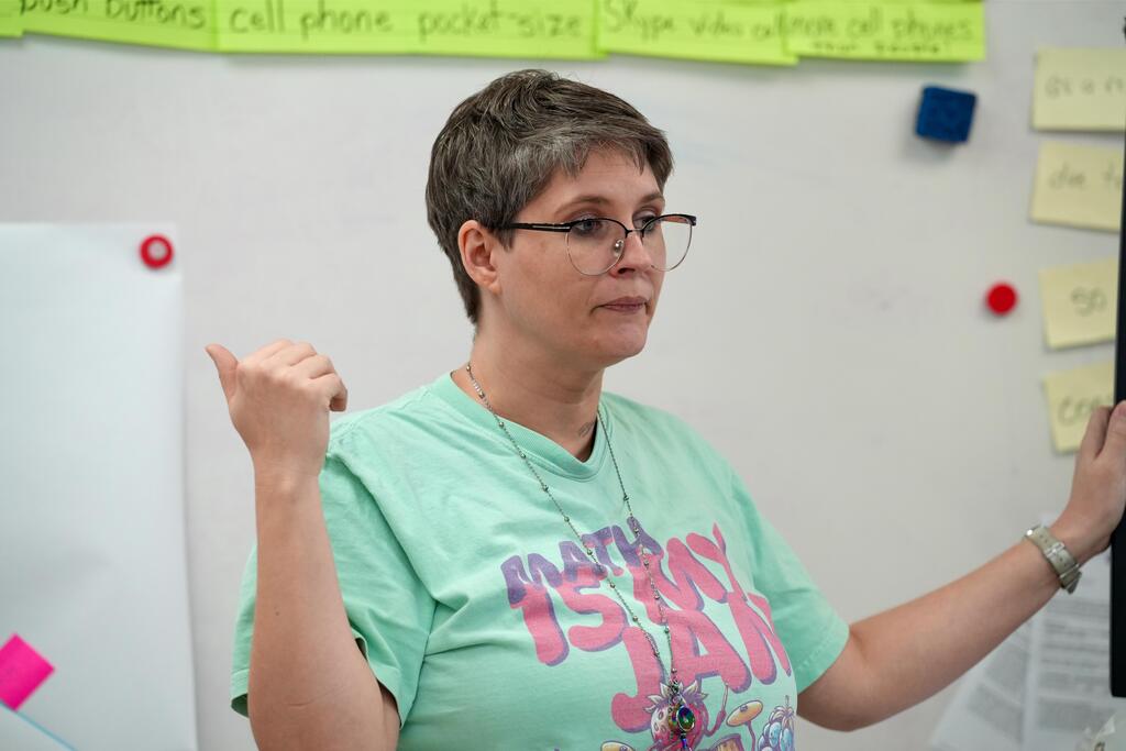 A person with glasses points to a screen while standing in a classroom. A white wall with colorful notes is behind.