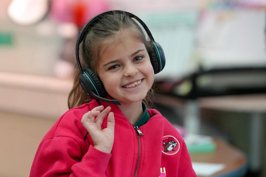 A young girl in a red jacket with a logo wears headphones, smiling, and waving in a classroom.
