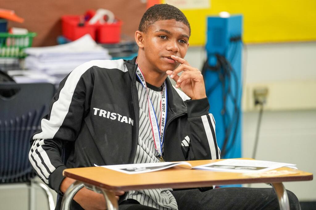 A student sits in a classroom, wearing a black and white jacket with "TRISTAN" on the back.