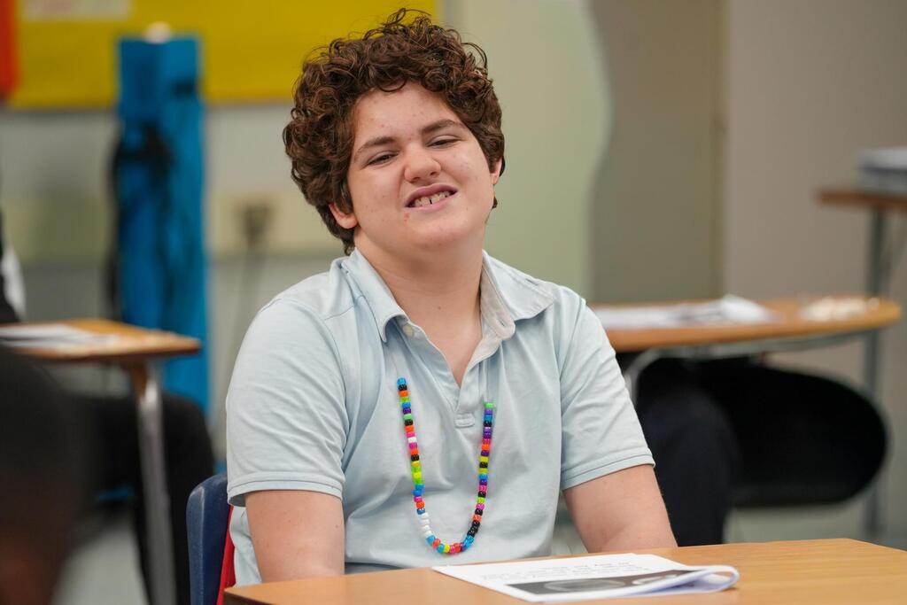 A person with curly hair smiles in a classroom, wearing a light blue shirt and colorful necklace.