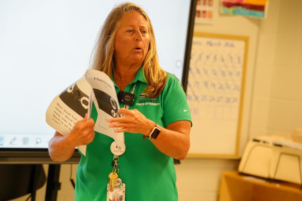 A woman in green stands in a classroom, holding a paper. Behind her is a projector screen and a bulletin board.