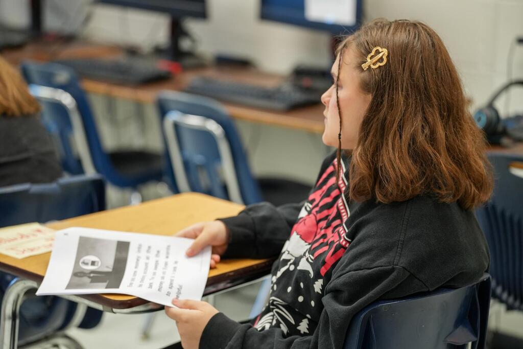 A student with a headphone reads a paper in a classroom with other students and computers.