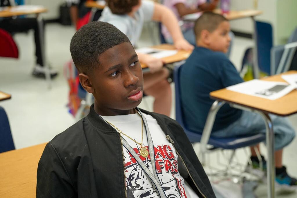A boy with a necklace and t-shirt sits at a desk in a classroom with other students.