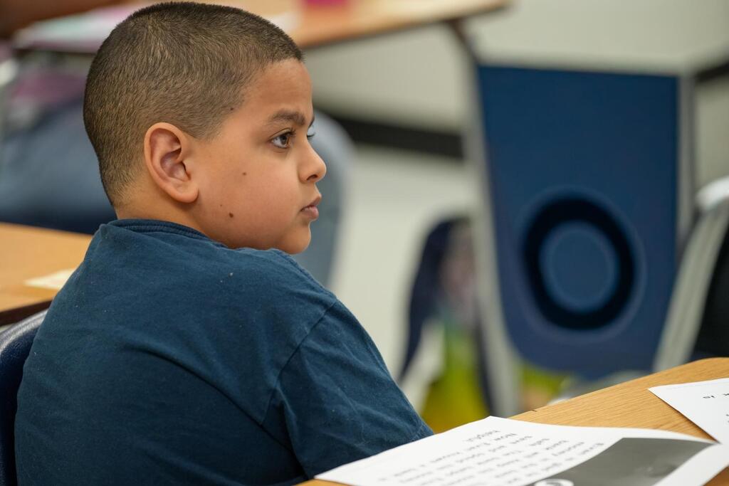 A young child sits at a desk in a classroom, facing sideways. Papers and a book are on the desk.