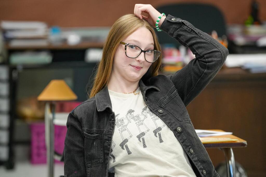 A young woman in glasses and a jacket poses in a classroom, with a desk and a lamp behind her.