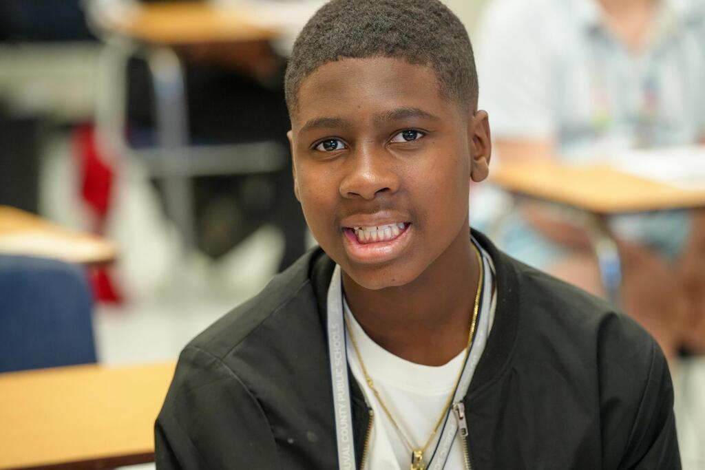 A young boy wearing a black jacket and necklace is smiling, seated in a classroom with desks and chairs.
