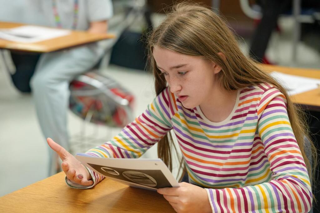 A girl in a colorful striped shirt sits at a desk in a classroom, reading a book.