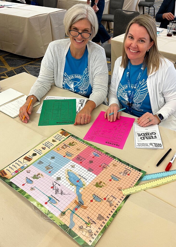 Two women seated at a table with a board game set up, one wearing glasses and a watch, both smiling.